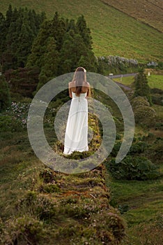 Young bride in white dress standing alone on grassy brickwall. Vie from behind. Background of romantic green hills