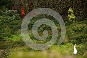 Young bride in white dress standing in field near grassy brickwall. Small woman figure in green fairy tale landscape