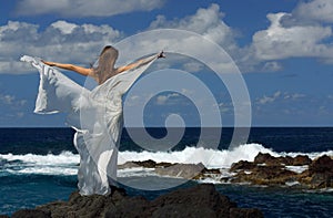 Young bride with white wings of wedding dress on rock sea shore. Back view. Ocean waves, splash of water