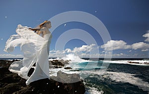 Young bride with white wings of wedding dress on rock sea shore. Side view. Ocean waves, splash of water