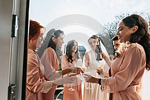 Young bridesmaids are having fun and smiling in pink silk robes drinking champagne at the bride's gazebo.