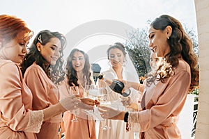 Young bridesmaids are having fun and smiling in pink silk robes drinking champagne at the bride's gazebo.