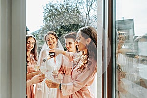 Young bridesmaids are having fun and smiling in pink silk robes drinking champagne at the bride's gazebo.