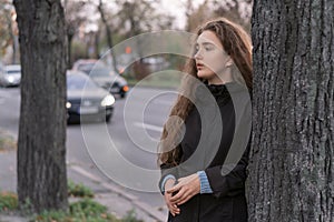 Young brooding woman stands by tree near highway on cloudy autumn day. Teen girl in coat on road background