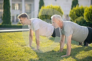Young brown-haired male and mature grey-haired male doing push ups on grass