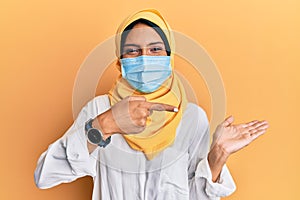 Young brunette arab woman wearing traditional islamic hijab and medical mask amazed and smiling to the camera while presenting