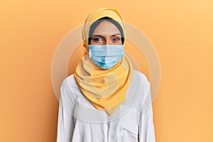 Young brunette arab woman wearing traditional islamic hijab and medical mask looking positive and happy standing and smiling with