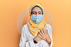 Young brunette arab woman wearing traditional islamic hijab and medical mask smiling with hands on chest with closed eyes and