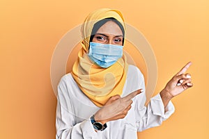 Young brunette arab woman wearing traditional islamic hijab and medical mask smiling and looking at the camera pointing with two