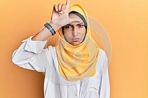 Young brunette arab woman wearing traditional islamic hijab scarf making fun of people with fingers on forehead doing loser