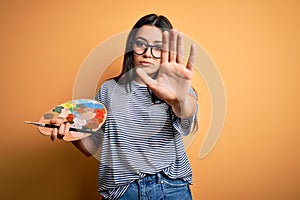 Young brunette artist woman painting holding painter brush and palette over yellow background with open hand doing stop sign with