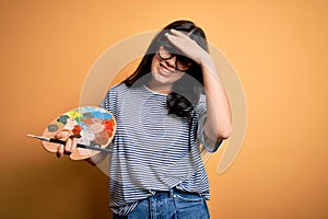 Young brunette artist woman painting holding painter brush and palette over yellow background stressed with hand on head, shocked