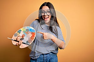 Young brunette artist woman painting holding painter brush and palette over yellow background very happy pointing with hand and