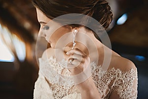 Young brunette beauty with wedding hairstyle in fashionable lace dress adjusting her earring and posing at interior