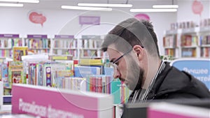 Young brunette black-haired man with glasses looking books on the bookshelves in the bookstore.