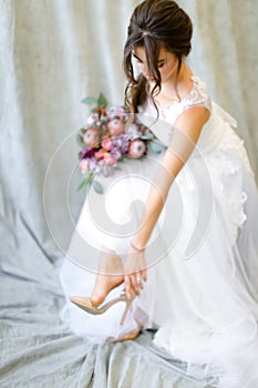 Young brunette bride with flowers putting on shoes at photo studio.
