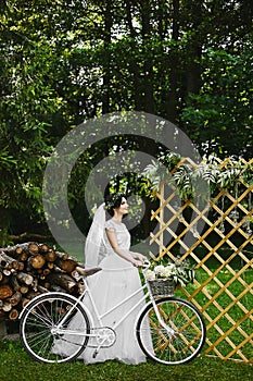 Young brunette bride in white fashionable dress with bicycle and with a bouquet of flowers in the summer garden