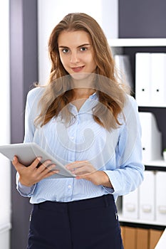 Young brunette business woman looks like a student girl at work in office. Girl standing straight with tablet pc