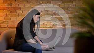 Young brunette caucasian girl in profile sitting on sofa and typing on laptop attentively in home atmosphere.