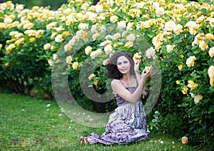 Young brunette Caucasian woman with curly hair sitting on green grass near yellow roses Bush in a garden, looking straight to the