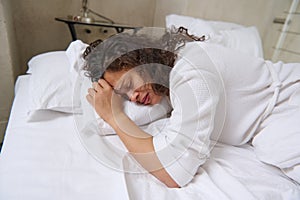 Young brunette crying and feeling depression, lying on white bed sheets in her bed in the home bedroom