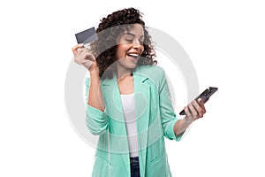 young brunette curly business woman in a jacket uses internet banking and a card in a smartphone