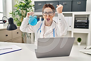Young brunette doctor woman holding alarm clock annoyed and frustrated shouting with anger, yelling crazy with anger and hand