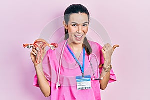 Young brunette doctor woman holding anatomical model of female genital organ pointing thumb up to the side smiling happy with open