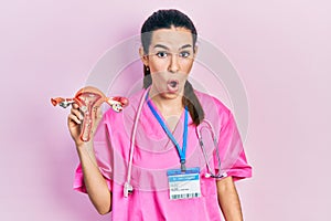 Young brunette doctor woman holding anatomical model of female genital organ scared and amazed with open mouth for surprise,