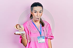 Young brunette doctor woman holding anatomical model of female uterus with fetus scared and amazed with open mouth for surprise,