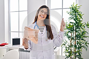 Young brunette doctor woman holding cervical neck collar smiling happy pointing with hand and finger to the side