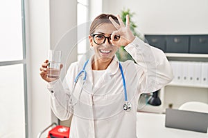 Young brunette doctor woman holding glass of water smiling happy doing ok sign with hand on eye looking through fingers