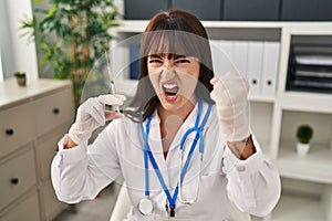 Young brunette doctor woman holding syringe annoyed and frustrated shouting with anger, yelling crazy with anger and hand raised