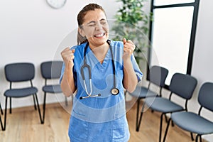 Young brunette doctor woman at waiting room very happy and excited doing winner gesture with arms raised, smiling and screaming