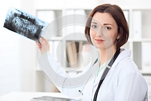 Young brunette female dentist sitting at the table and examines dental X-ray