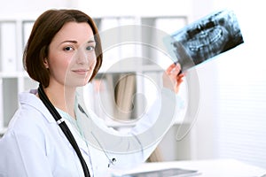 Young brunette female dentist sitting at the table and examines dental X-ray