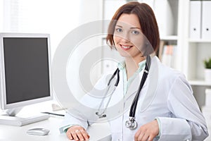 Young brunette female doctor sitting at a desk and working on the computer at the hospital office.