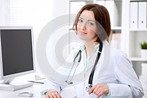 Young brunette female doctor sitting at a desk and working on the computer at the hospital office.