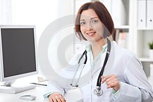 Young brunette female doctor sitting at a desk and working on the computer at the hospital office.