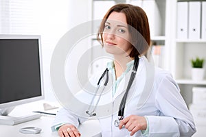 Young brunette female doctor sitting at a desk and working on the computer at the hospital office.