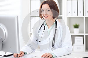 Young brunette female doctor sitting at a desk and working on the computer at the hospital office.