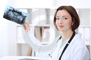 Young brunette female doctor sitting at the table and considers x-ray picture of teeth