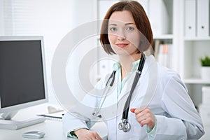 Young brunette female doctor sitting at the table and working with computer at hospital office