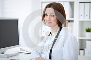 Young brunette female doctor sitting at the table and working with computer at hospital office