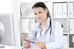 Young brunette female doctor sitting at the table and working by computer at hospital office.