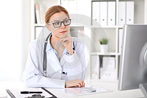 Young brunette female doctor sitting at the table and working by computer at hospital office.