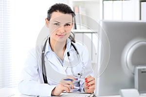 Young brunette female doctor sitting at the table and working by computer at hospital office.