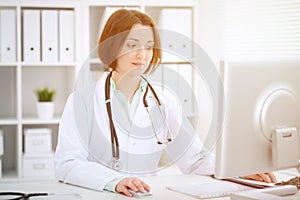 Young brunette female doctor sitting at the table and working with computer at hospital office