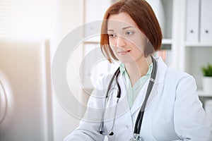 Young brunette female doctor sitting at the table and working with computer at hospital office