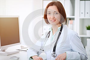 Young brunette female doctor sitting at the table and working with computer at hospital office